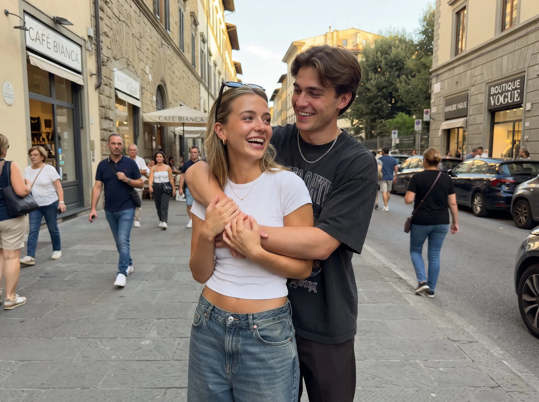 A young couple laughing on a city street in a back hug, his arms wrapped around her waist and her hands on his arms