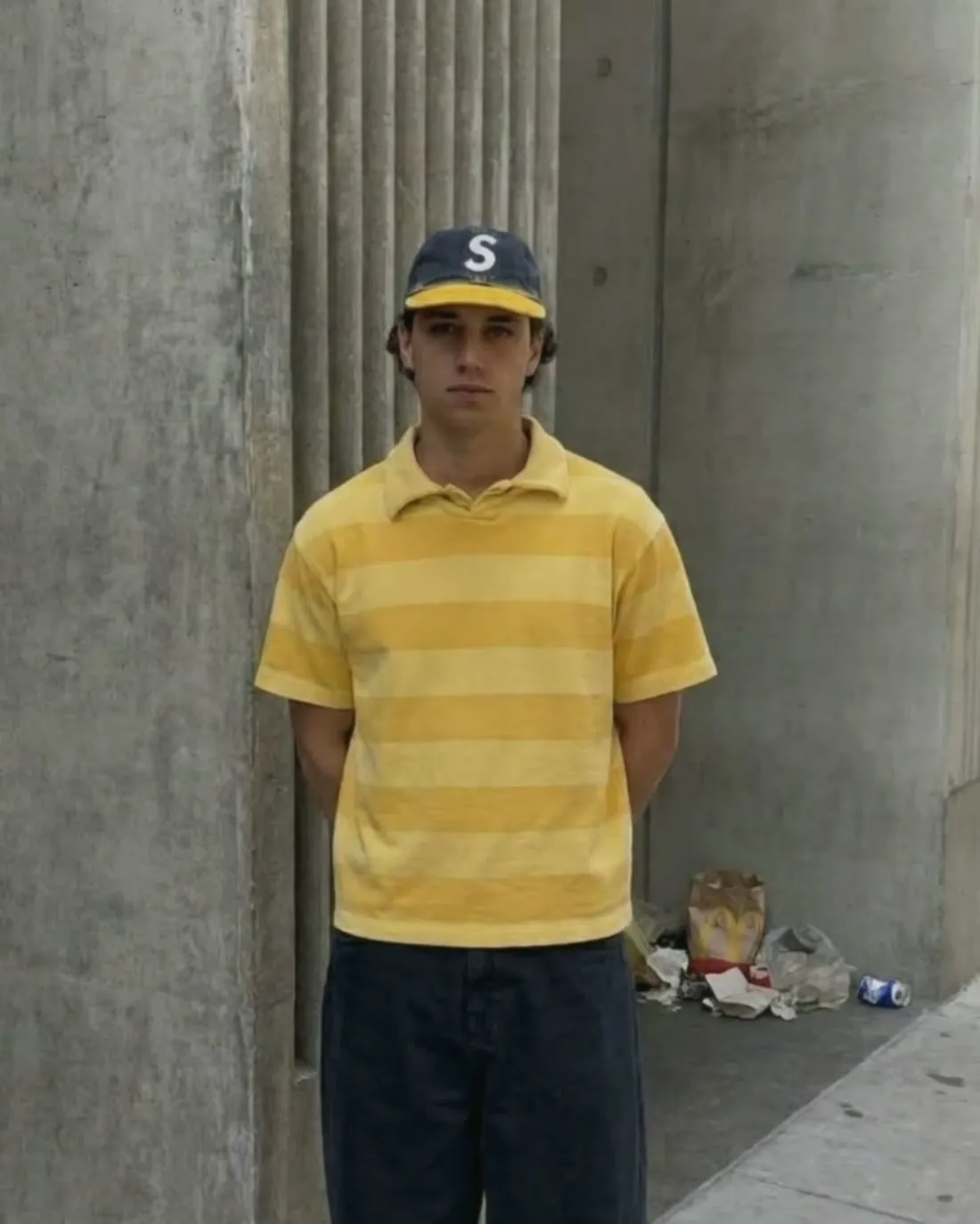 Man in a yellow striped shirt and cap standing stiffly against a concrete wall with his hands behind his back